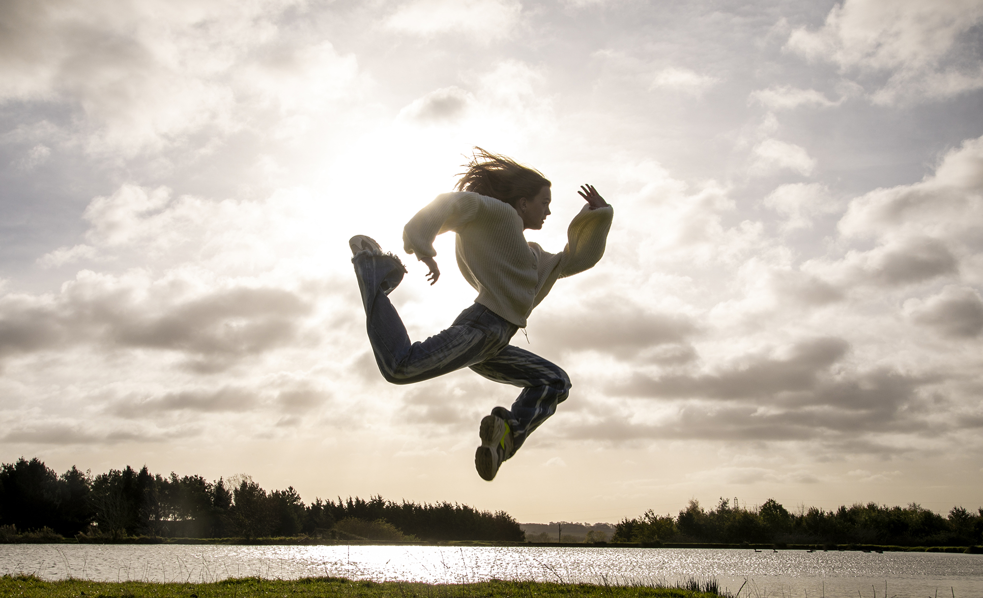 white female dancer jumping in the air in a running shape in front of golden clouded sky wearing casual clothes